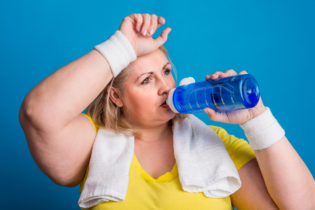 Portrait of a tired overweight woman in studio on a blue background, drinking water.の写真素材