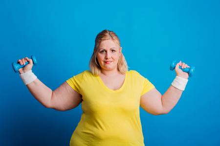 Portrait of a serious overweight woman with dumbbells in studio on a blue background.の写真素材