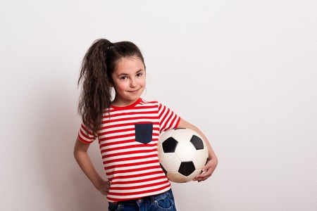 A small girl with a soccer ball and striped T-shirt standing in a studio.の写真素材