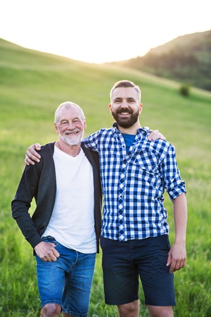 A portrait of an adult hipster son with senior father in nature at sunset.