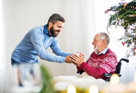 A hipster man giving drink to his senior father in wheelchair at Christmas time.