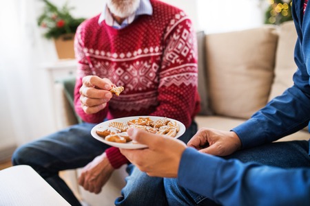 Unrecognizable senior father and adult son sitting on a sofa at Christmas time, eating biscuits.