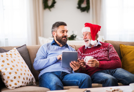 A senior father and adult son with tablet sitting on a sofa at home at Christmas time.の写真素材
