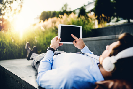 A businessman with tablet and headphones, lying on steps outdoors at sunset.の写真素材
