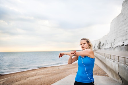 Young sporty woman runner with earphones standing on the beach outside, stretching.の写真素材