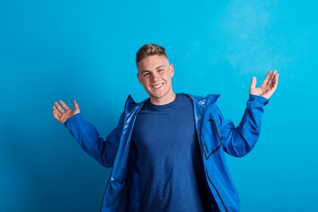 Portrait of a young man with blue anorak in a studio, standing against blue background.の写真素材