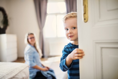 A toddler boy with young mother in the background inside in a bedroom.