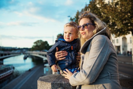 Young mother with a toddler son standing outdoors by the river in city.の写真素材
