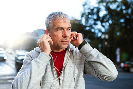 A portrait of an active mature man with earphones standing outdoors in city.の写真素材