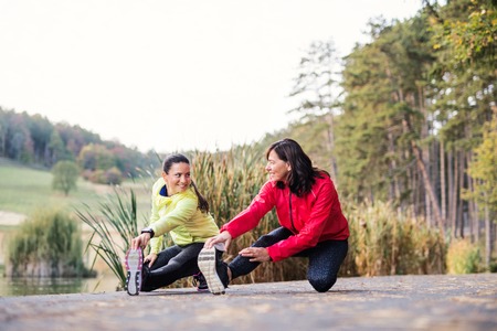 Two female runners stretching legs outdoors in park in autumn nature.