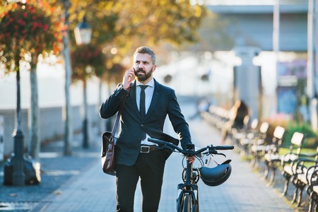 Businessman commuter with bicycle walking home from work in city, using smartphone.の写真素材