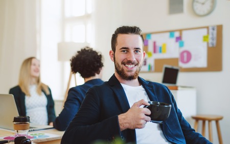 A portrait of young businessman with coffee and colleagues in a modern office.