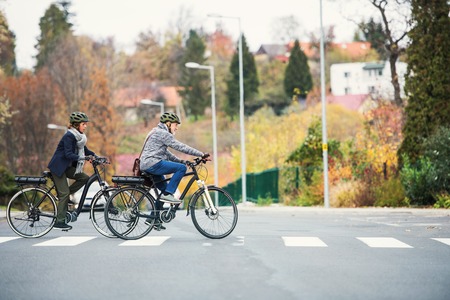 Active senior couple with electrobikes outdoors crossing a road in town.の写真素材