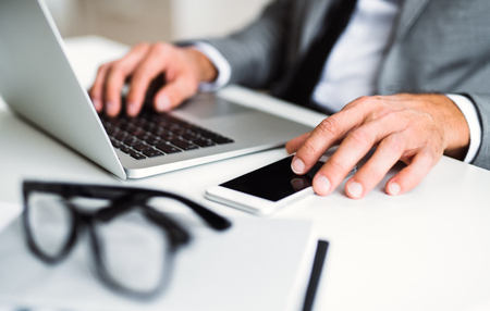 A midsection of businessman with smartphone and laptop sitting at the table, working.の写真素材