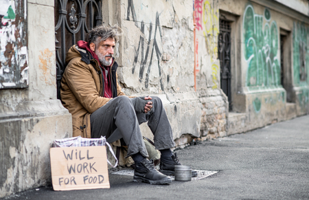 Homeless beggar man sitting outdoors in city asking for money donation.