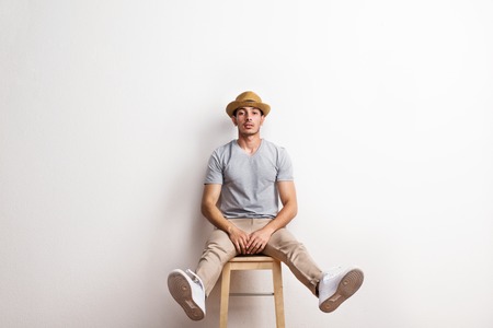 A confident hispanic young man with hat sitting on a stool in a studio.の写真素材