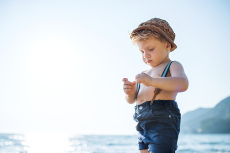 A small toddler boy with hat and shorts standing on beach on summer holiday.の写真素材