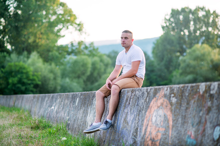 Young lonely man sitting on a stone wall in sunny summer nature.の写真素材
