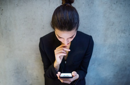A top view of young business woman with smartphone standing against concrete wall in office.の写真素材