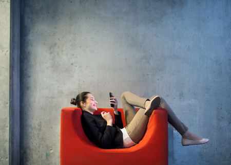 A young businesswoman with smartphone sitting on red armchair in office.の写真素材