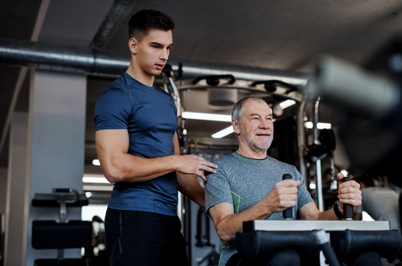 A senior man with a young trainer doing strength workout exercise in gym.の写真素材