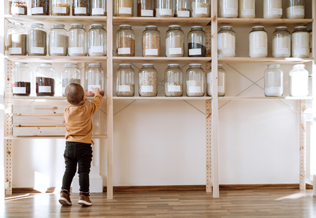 A rear view of small toddler boy standing by shelf with glass jars in zero waste shop.の写真素材