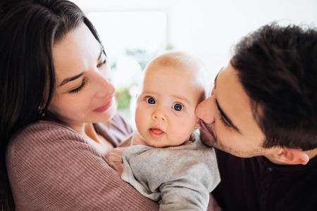 A close-up portrait of young couple with a baby indoors at home.