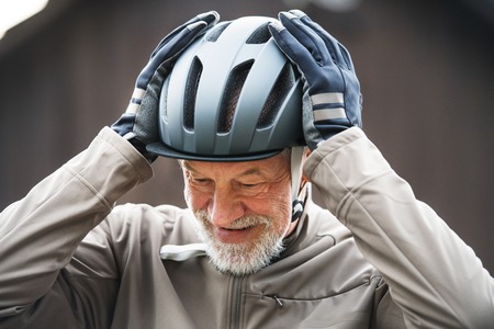 An active senior man standing outdoors in town, putting on a bike helmet.