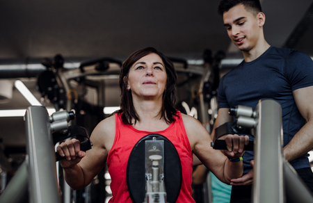 A female senior with a young trainer doing strength workout exercise in gym.の写真素材