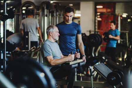 A senior man with a young trainer doing strength workout exercise in gym.の写真素材