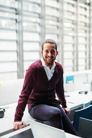 A young businessman sitting on a desk in office, looking at camera.の写真素材