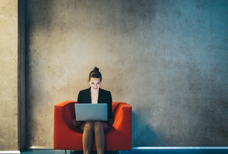 A young businesswoman with laptop sitting on red armchair in office. Copy space.の写真素材