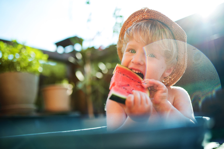 Small boy with a hat in bath outdoors in garden in summer, eating watermelon.の写真素材