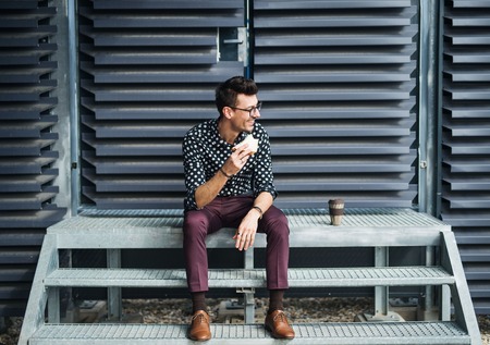 A young businessman with coffee and sandwich sitting outdoors, having a snack.の写真素材