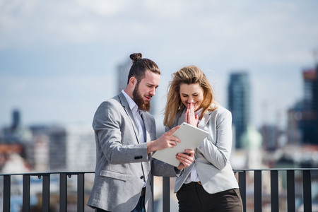Two young business people with tablet standing on a terrace outside office, working.