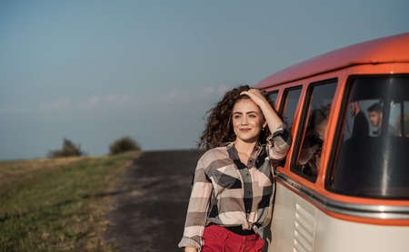 A young girl on a roadtrip through countryside, leaning on a minivan.