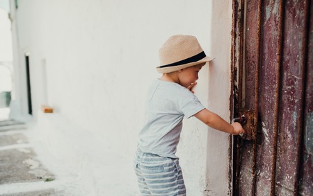 A small toddler boy with hat standing by a house in town on summer holiday.の写真素材