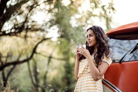 A young girl with coffee by a car on a roadtrip through countryside.