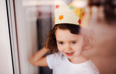 Portrait of small girl with crown at home, looking out of a window. Shot through glass.