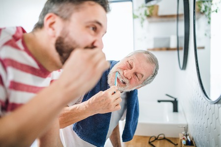 An adult hipster son and senior father brushing teeth indoors at home.