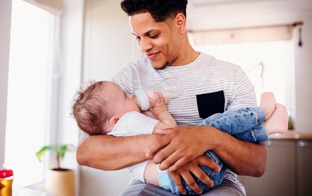 A portrait of father bottle feeding a small toddler son indoors at home.