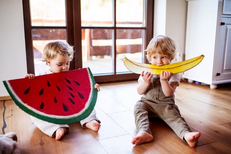 Two toddler children playing with large toy fruit indoors at home.の写真素材