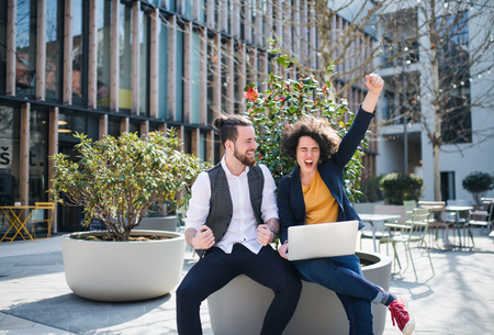 Young businesspeople with laptop outdoors in courtyard, expressing excitement.の写真素材