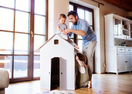 Two toddler children with father playing with paper house indoors at home.の写真素材