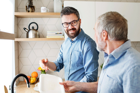 Adult hipster son and senior father indoors in kitchen at home, washing dishes.の写真素材