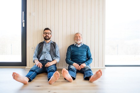 A portrait of adult hipster son and senior father sitting on floor indoors at home.の写真素材