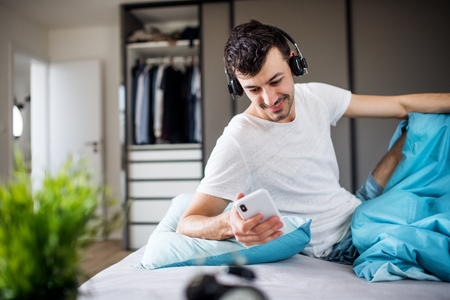 Young man with headphones and smartphone in bed at home, listening to music.の写真素材