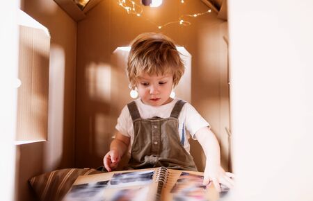 A toddler boy playing indoors in cardboard house at home, looking at photo album.の写真素材