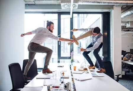 Two young businessmen with swords in an office, having fun. A competition concept.の写真素材