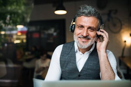Mature man with headphones at the table in a cafe, using laptop.